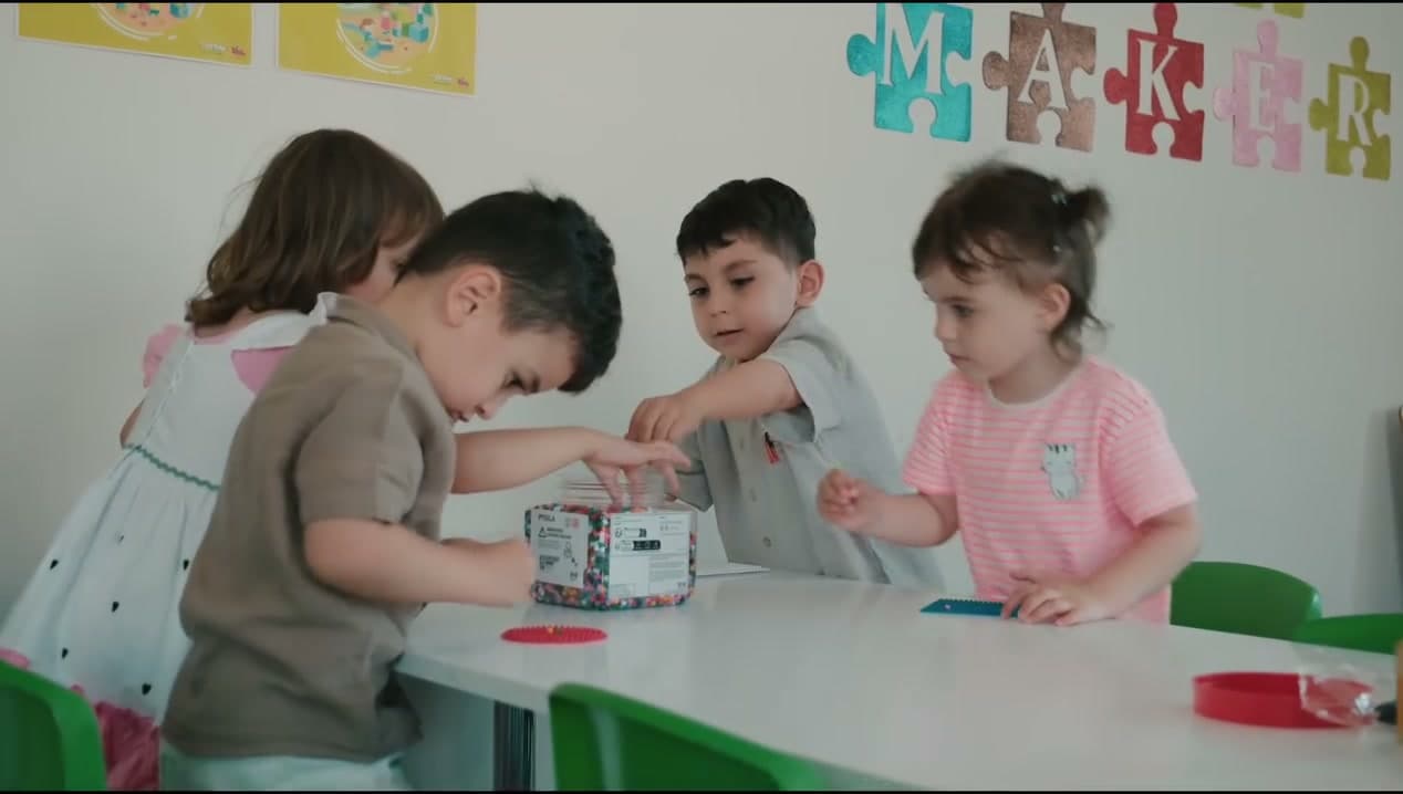 MAKER yazılı atölye sınıfında masa başında boncuk etkinliği yapan dört çocuk